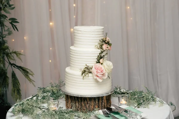 Beyaz düğün pastası, ışıkların arka planında duran ahşap bir tezgahta çiçeklerle süslenmiş. Yeni evliler için lezzetli, lüks, çok katlı bir pasta. Pastanın üzerindeki güller