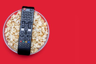 Popcorn in a glass bowl and a TV remote control on a red background. The concept of watching movies or series with a crispy snack in the form of popcorn. Top view of popcorn. Free space for text