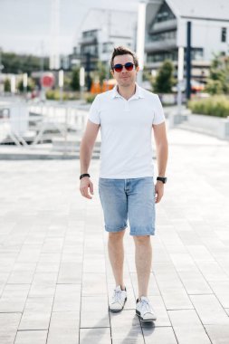 Şort, beyaz tişört ve güneş gözlüğü giyen şık bir adam. Şık bir saç stili ve şık aksesuarları olan bir adam sokakta yürüyor.