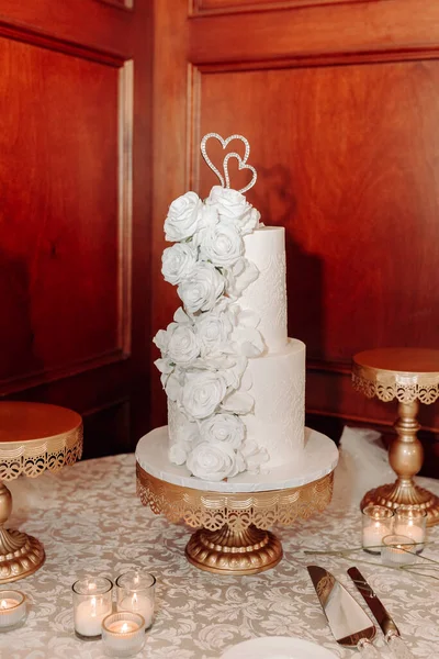 The white wedding cake is decorated with roses and a figurine in the form of two hearts. Delicious luxurious tiered honeymoon cake. Traditional wedding cake. Chic white cake on a vertical image