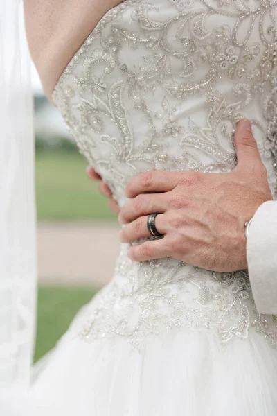 The groom hugs the bride dressed in a white wedding dress. Close-up of the groom\'s hands holding the bride\'s waist. Wedding day and hugs close up. The groom has a black wedding ring on his finger