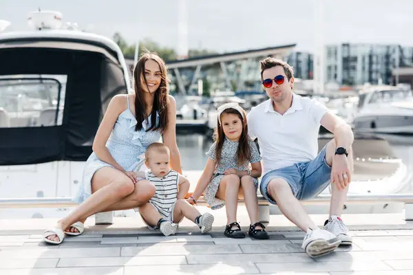 A family of four sits against the backdrop of yachts in the harbor. Mom and dad together with their little son and daughter. Happy family spending free time outdoors in the evening