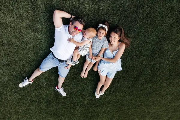 A happy family of four lies on the green lawn and smiles. Dad, mom, brother and sister are lying on the manicured lawn. Happy parents and their little children having fun