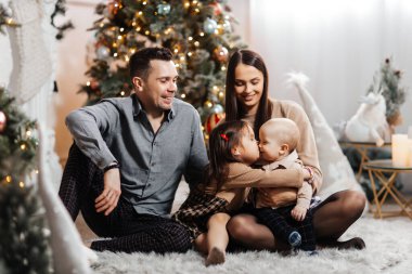 Mutlu aile, yeni yıl ağacı ve Noel dekorunun arka planında eğleniyor. Genç baba ve mutlu anne, oğlu ve kızı gülümseyip yeni yılın tadını çıkarıyorlar. Abla abiyi öptü