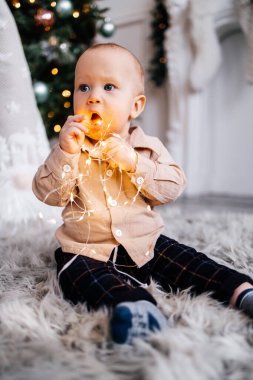 Bir çocuk Noel ağacının arka planında çelenkle oynuyor. Sevimli bir çocuk elinde parlak bir çelenk tutuyor. Noel atmosferi ve tatlı küçük çocuk