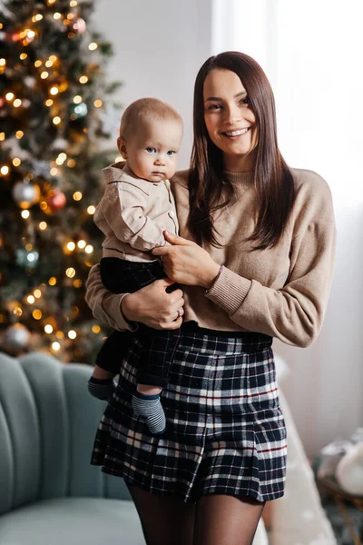 A happy young mother holds her little son in her arms against the backdrop of a beautifully decorated New Year tree. Christmas atmosphere and happy family. Beautiful portrait of mother and son