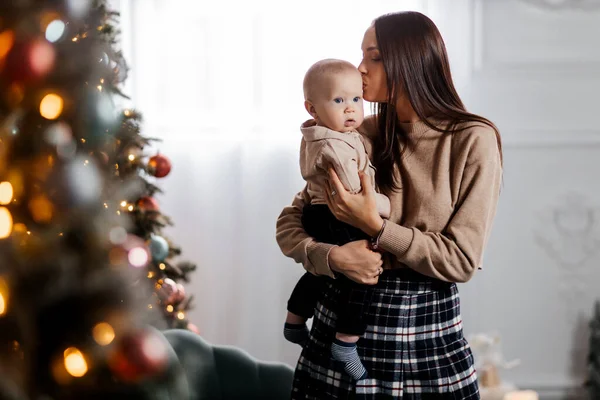 A happy young mother holds her little son in her arms against the backdrop of a beautifully decorated New Year tree and kisses him. Christmas family atmosphere. Beautiful portrait of mother and son