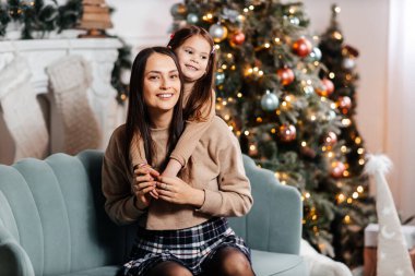 Genç ve mutlu bir anne ve kızı güzel dekore edilmiş bir Noel ağacının önünde sarılıyorlar. Noel atmosferi ve mutlu bir aile. Anne ve kızın güzel portresi