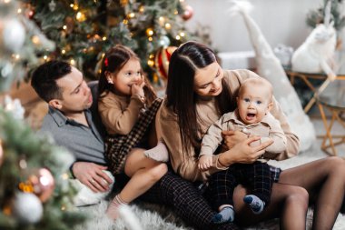 Mutlu bir aile, yeni yıl ağacı ve Noel dekoruna karşı birlikte eğleniyor. Genç baba ve mutlu anne, oğlu ve kızı gülümseyip yeni yılın tadını çıkarıyorlar.
