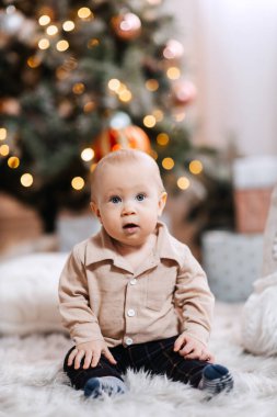 Bir çocuk, çok renkli toplarla süslenmiş bir Noel ağacının arka planında çelenkle oynuyor. Sevimli bir çocuk elinde parlak bir çelenk tutuyor. Noel atmosferi ve tatlı küçük çocuk