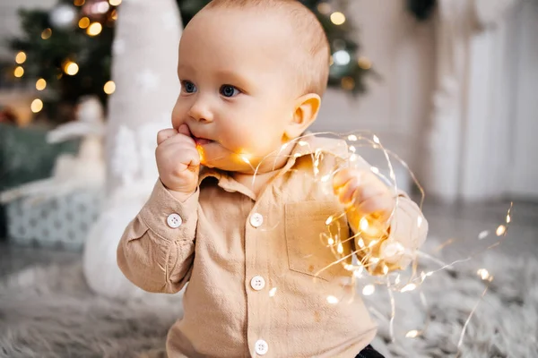 Çocuk bir Noel ağacının arka planında çelenkle oynuyor. Sevimli bir çocuk elinde parlak bir çelenk tutuyor. Noel atmosferi ve bej gömlekli şirin çocuk.
