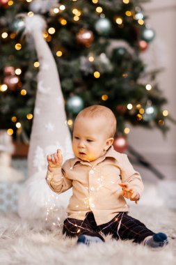Çocuk bir Noel ağacının arka planında çelenkle oynuyor. Sevimli bir çocuk elinde parlak bir çelenk tutuyor. Noel atmosferi ve mavi gözlü sevimli küçük çocuk