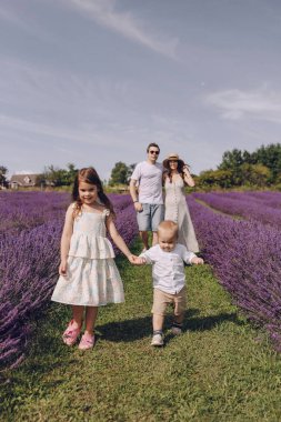 Genç ve mutlu bir aile, çocuklarıyla birlikte çiçek açan lavanta tarlasında yürüyor. Küçük erkek kardeş kız kardeşinin elini tutuyor. Genç bir aile lavanta tarlasında yürüyor. Mutlu çocukluk ve annelik