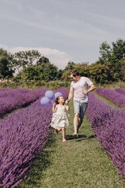 Baba ve kızı mor lavanta tarlasında. Baba ve kızı el ele tutuşup sahada koşarken eğleniyorlar. Küçük bir kız elinde balonlar tutuyor. Aile çiçek açan bir çayırda yürüyor