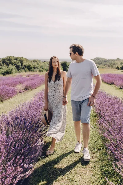 A couple in love walks through a lavender field. A woman in a dress and a straw hat with a man hold hands and walk through a purple lavender field. Romantic relationship