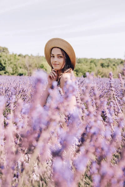 Beautiful young woman in the middle of a blooming lavender field. Young brunette in a straw hat at a lavender farm in Provence. Beautiful purple lavender field. Portrait of a cute woman posing