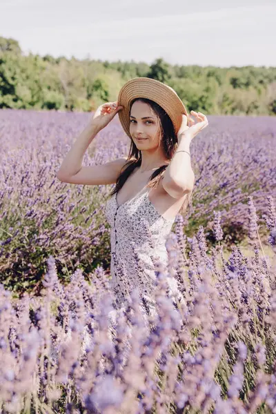 Beautiful young woman in the middle of a blooming lavender field. Young brunette in a straw hat at a lavender farm in Provence. Beautiful purple lavender field. Vertical portrait of a posing woman
