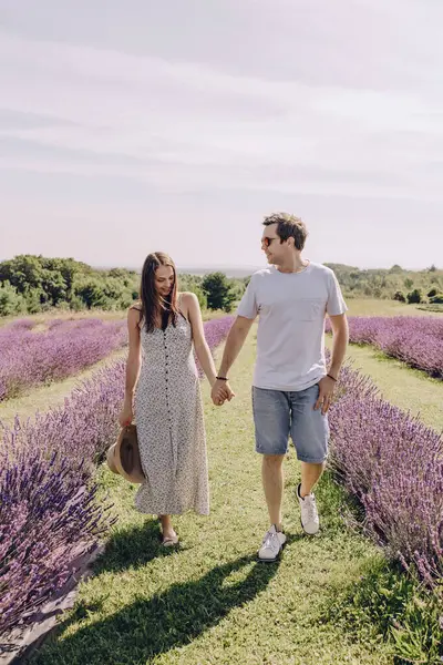 A couple in love walks through a purple lavender field. A woman in a dress and a straw hat with a man hold hands and walk through a purple lavender meadow. Romantic relationship. Unity with nature