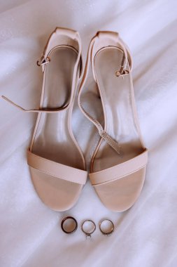 Gelin için düğün ayakkabıları. Gelin için zarif krem rengi sandaletler alyansların yanındaki beyaz bir tül üzerinde duruyor. Gelinin aksesuarları. Düğün günü.