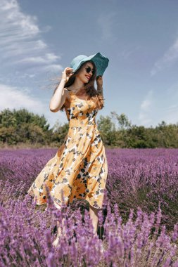 Genç bir kadın çiçek açan lavanta tarlasında yürüyor. Yazlık şapkalı, elbiseli ve güneş gözlüklü mutlu kızıl kadın gülümsüyor. Lavanta çayırında yaz yürüyüşü