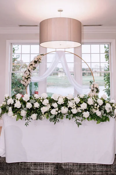 A garland of white roses on a table in a restaurant, behind a golden arch with delicate roses and tulle. A large window in the background. Wedding decorations, flower arrangement of roses.