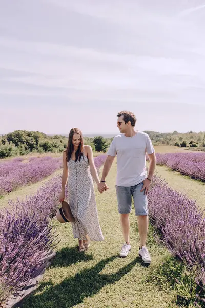 A couple in love walks through a purple lavender field. A woman in a dress and a straw hat with a man hold hands and walk through a lavender meadow. Romantic relationship between a man and a woman