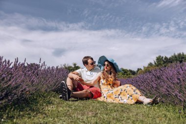 Lavanta tarlasında çimlerde oturan mutlu adam ve kadın. Kadın, adama yaslanıyor, adam da ona bakıyor. Genç bir çift lavanta çayırında nazikçe el ele oturuyor..