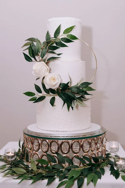 A white wedding cake is decorated with a wreath of fresh roses and green leaves on a beautiful golden plate. A three-tiered white cake stands on a light background, candles and green leaves around