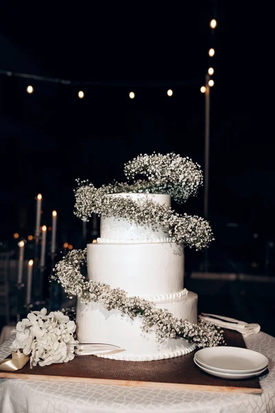 Three-tier wedding cake with white cream decorated with small white flowers. A beautiful white cake decorated with fresh white flowers stands outside at night. White holiday cake.