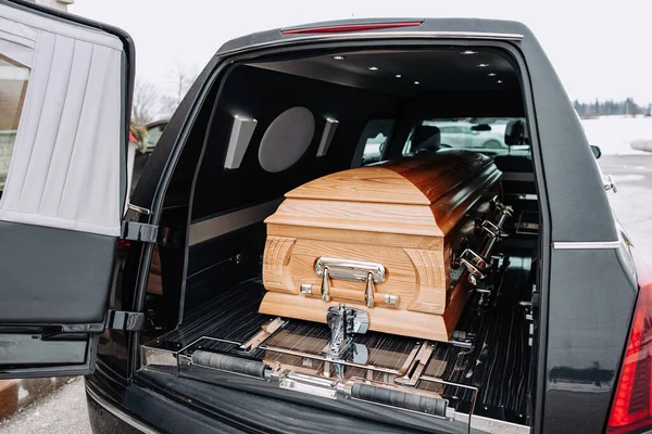A coffin made of light wood stands in the trunk of a black hearse. Funeral and farewell ceremony. Closeup photo of a funeral casket.