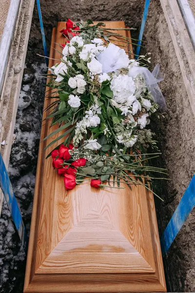 A wooden coffin in a grave. A coffin with white and red flowers on it is lowered into a dug grave. Funeral ceremony at the cemetery. Farewell ceremony and burial. Close-up photo.