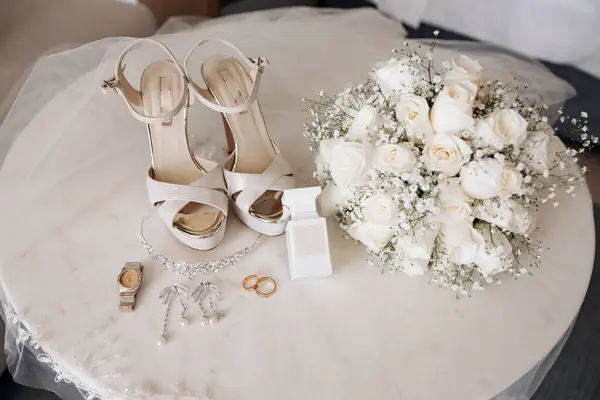 Wedding accessories. The bride\'s bouquet of white roses lies on the table, high-heeled shoes, two gold wedding rings, earrings, a diadem, a watch, and perfume are nearby. Wedding details.
