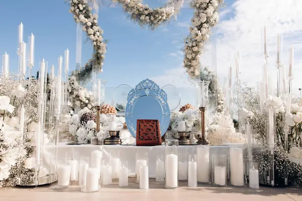Beautiful wedding decorations in white colors outside according to Muslim traditions. Wedding arch made of white flowers, white roses, white candles in glass candle holders, mirror, Koran.