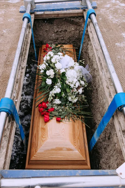 A wooden coffin with white and red flowers on it in a grave. A coffin with flowers is lowered into a dug grave. Funeral ceremony at the cemetery in winter. Farewell ceremony and burial.
