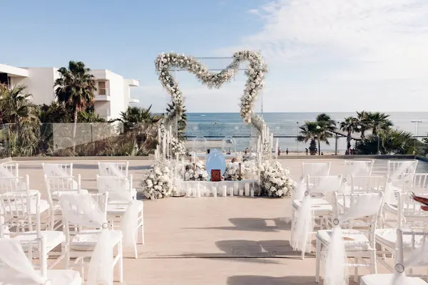 Wedding ceremony in the open air against the background of the ocean. In the center is a beautiful wedding arch made of white flowers, white candles, the Koran. Decorated white chairs for guests.