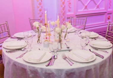 Masada boş çatal bıçaklar, mumlar ve çiçekler olsun. Beyaz masa örtülü yuvarlak servis masası ve davetsiz misafirler için boş tabaklar, kutlama, akşam yemeği.