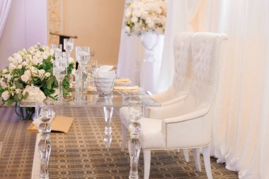A wedding table and two white chairs for the bride and groom. The table set for the newlyweds is decorated with white flowers and candles. The hall is decorated with flowers for the wedding