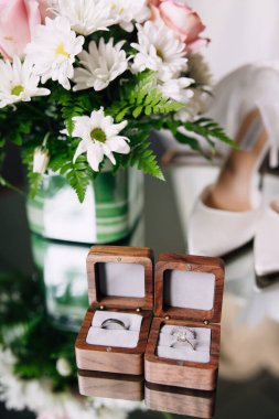 Wedding rings of the bride and groom in a wooden box close-up. Bride's bouquet and bride's elegant wedding shoes in the background. Two rings with stones of the bride and a ring of the groom