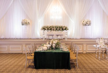Büyük bir düğün salonu. Düğün masası, sahnedeki yeni evliler için çiçekler ve iki beyaz sandalyeyle süslenmiştir. Salon bir düğün ve davetsiz bir şenlik masası için dekore edildi.