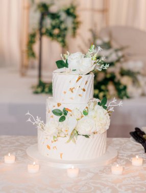 Çiçekler ve altınla süslenmiş güzel beyaz pasta. Mumlarla süslenmiş düğün pastası ve masanın üzerinde beyaz güller var. Pasta şeklinde güzel bir şekerleme.