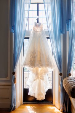 Pencerede asılı beyaz gelinlik. Güzel bir odada direğe asılı lüks bir gelinlik. Gelinin elbisesinin dikey fotoğrafı.