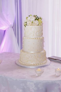 Gelin ve damadın düğün yemeği için güllerle süslenmiş üç katlı beyaz pasta. Lokantadaki bir masada lüks bir doğum günü pastası.