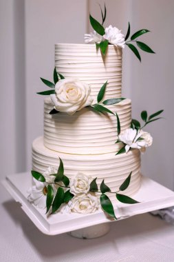 Çiçeklerle süslenmiş beyaz düğün pastası masanın üzerinde duruyor. Gelin ve damadın pastası beyaz güllerle süslenmiştir. Geleneksel düğün pastası. Kutlama partisi için güzel bir pasta.