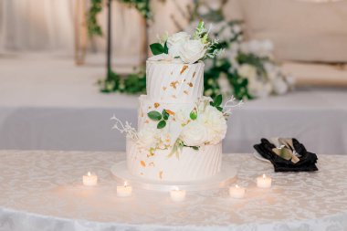 Çiçekler ve yenilebilir altınla süslenmiş güzel beyaz pasta. Mumlarla süslenmiş düğün pastası ve masanın üzerinde beyaz güller var. Pasta şeklinde güzel bir şekerleme.