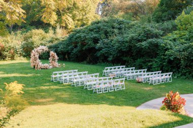 Bir sıra beyaz tahta sandalye ve açık havada güzel yuvarlak bir düğün kemeri. Kurumuş çiçeklerle süslenmiş yuvarlak düğün kemeri çimenlerin üzerinde taze güller ve dışarıda misafirler için boş beyaz sandalyeler..