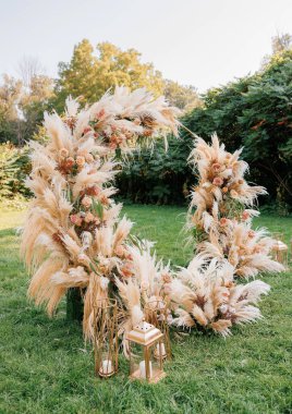 Boho tarzında güzel, yuvarlak bir düğün kemeri. Çimlerin üzerinde kurumuş çiçekler ve taze güllerle süslenmiş yuvarlak bir düğün kemeri. Düğün kemeri kırsal tarzda dekore edilmiş.