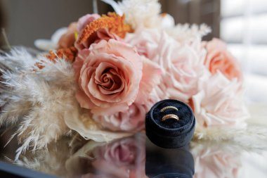 Bir kutuda iki gelin için altın alyanslar. Siyah bir kutuda taşlı iki alyans ve arkasında pembe güller ve kurumuş çiçekler olan güzel bir düğün buketi..