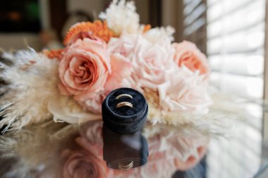 Bir kutuda iki kadın için iki alyans ve arkasında pembe güller ve kurumuş çiçekler olan güzel bir düğün buketi. Evlilik yüzükleri ve gelinin buketi masaya yansıyor..