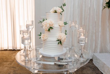 Üç katlı zarif düğün pastası ve taze beyaz güller. Kremalı vanilyalı bir düğün pastası cam bir masanın üzerinde durur ve etrafında su olan bardaklarda mumlar. Düğün davetlileri için şenlikli pasta.