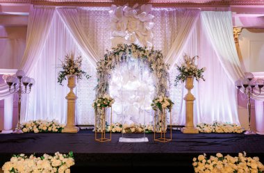 Beyaz çiçeklerle süslenmiş cam kürsü sahnesi. Arkasında çiçek kemeri ve gür beyaz perdeleri olan sahne. Zarif dekore edilmiş sahne, pembe gece ışıkları düğün ve törenler için mükemmel.